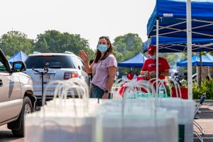 woman waving to camera in front of bags of to-go food at outdoor event